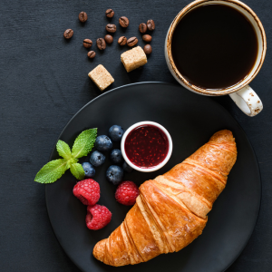 A delicious looking croissant on a dark plate surrounded by raspberries, jam and a dark coffee.