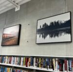 Two photographs by Cheryl Phillips hanging on a cement wall above bookshelves. One is a black and white of trees reflecting over water. The second shows a length of railroad cars on foggy day.