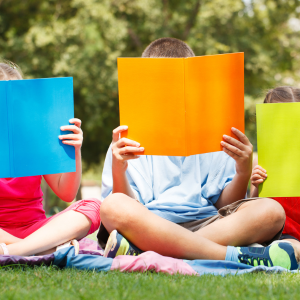 Three children sitting on the grass with books in front of their faces reading.