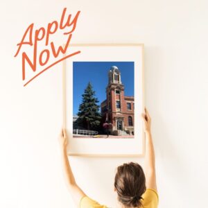 A person handing a photo of the library onto a wall with the words "Apply Now" written beside.