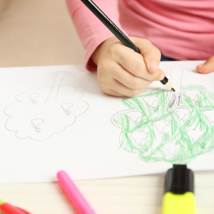 Child's hand sketching an apple tree with colouring material laying on the table.