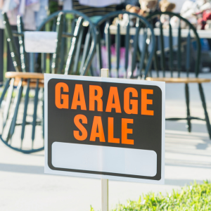 Garage Sale sign in a front yard with chairs, clothing and various items on a table in the background.