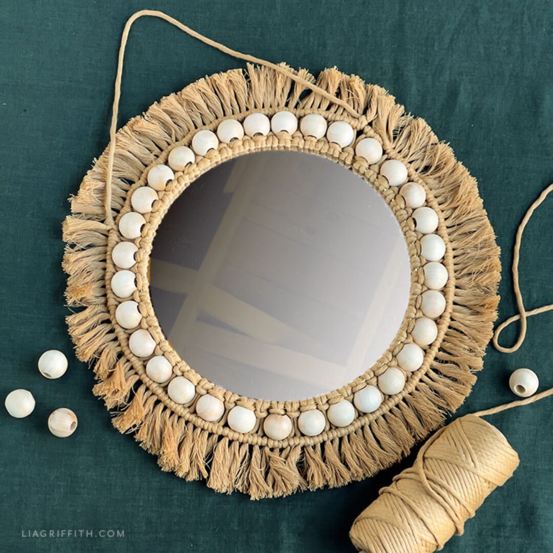 A round mirror surrounded by natural cotton rope tied in macrame knots. Laying on a dark blue-green linen tablecloth.