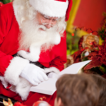 Santa sitting in front of a Christmas tree reading a book to a child.