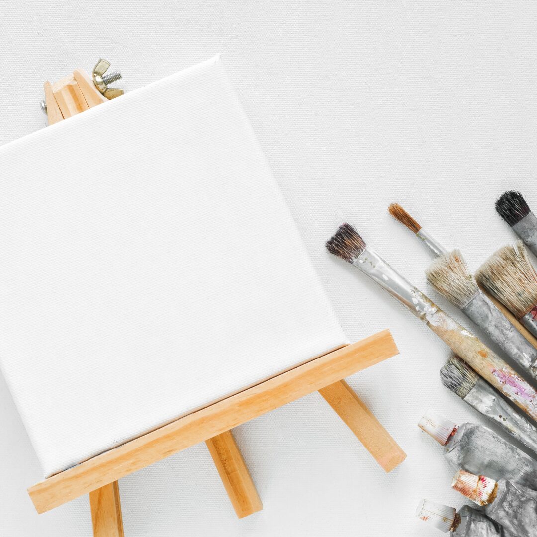 Blank mini art canvas on an easel with painting brushes and paint tubes lying beside it.