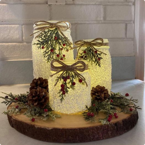 Three jars light by candles with a frosty coating. All send on a wood slab with holly and pinecones.