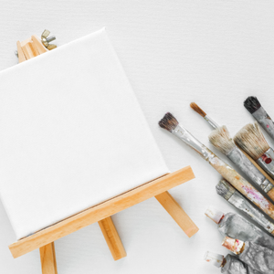 Small white unpainted canvas with paint brushes and paint beside it on a table top.