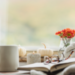 reading glasses on top of an open book with white pumpkins, a candle, orange flowers, and a cup of tea.