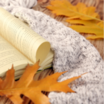 a book open on a wood table with a knit blanket and orange leaves