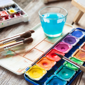 A palette of watercolour paints, a cup filled with ocean blue paint soaked water and used paintbrushes.